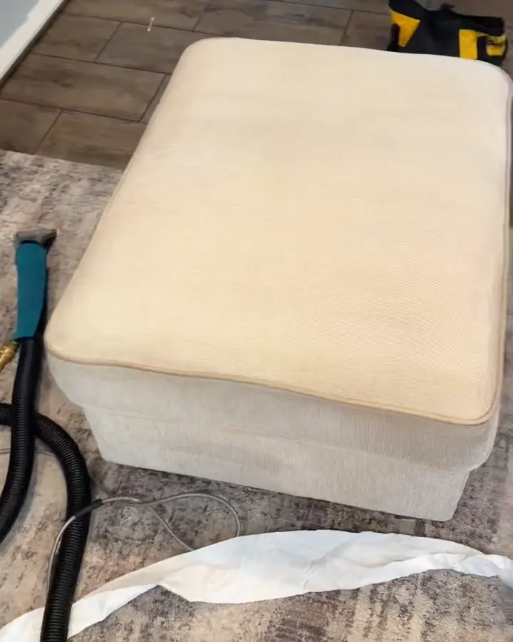 The same cream ottoman after cleaning, seen from above: the orange stain is gone and the top is still damp; the extraction wand and hose lie beside it.
