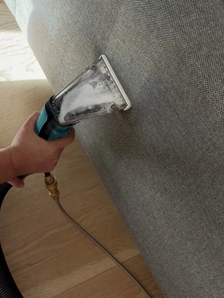 A clear extraction head pulling foamy dirty water out of the back of a gray sofa.