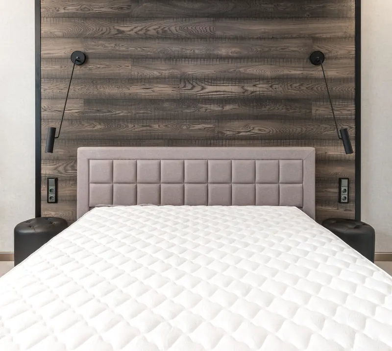 Colchón blanco acolchado sin sábanas sobre una cama con cabecera tapizada y pared de madera oscura.